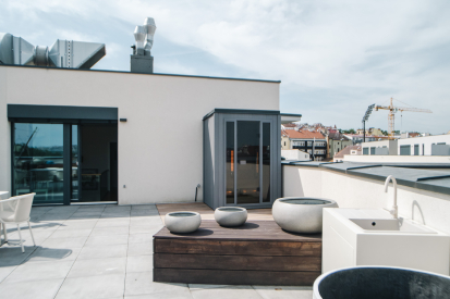 Venkovní infrasauna na střešní terase s výhledem Pohled na moderní střešní terasu s volně stojící prosklenou infrasaunou. V popředí jsou cementové květináče a venkovní dřez, v pozadí výhled na město.